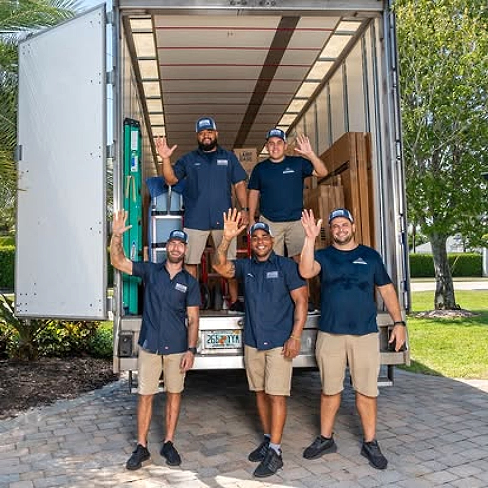 Credo Enterprise moving crew in navy uniforms waving beside an open truck loaded with boxes, moving blankets, and wooden crating supplies on a sunny Florida driveway